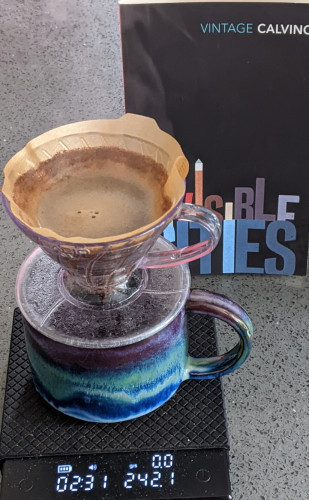 A coffee dripper sat on top of a beautiful mug swirled horizontally with bands of green and blue, which is on top of a black set of scales. Behind them is a copy of Invisible Cities by Italo Calvino