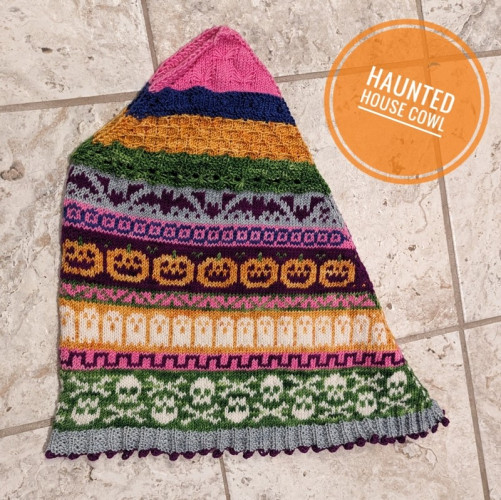 A picture of my finished knitted Haunted House cowl, which features a  number of different colourwork Halloween motifs including bats, jack-o-lanterns, ghosts and skull and crossbones. The whole piece is a strange trapezoidal shape when laid flat but will look somewhat like a triangle shawl worn around the neck.