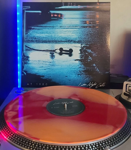 A Red & Orange Swirl vinyl record sits on a turntable. Behind the turntable, a vinyl album outer sleeve is displayed. The front cover shows a payphone handset laying in a wet street at night with it's cord still attached