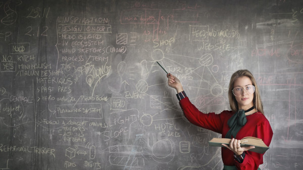 A tutor is standing bottom right of the photo, in front of a large blackboard. She is holding a book in her left hand, whilst looking towards her class, and pointing towards the blackboard with her right hand