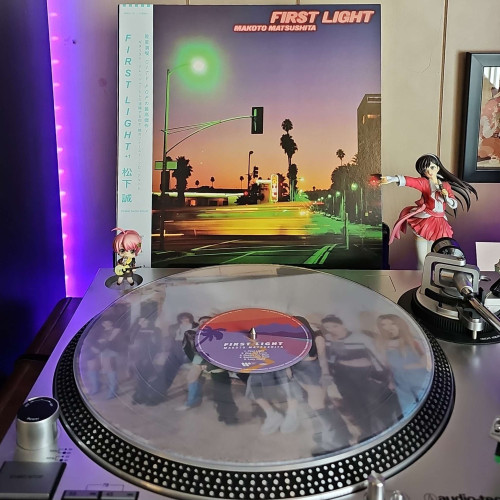 A clear vinyl record sits on a turntable. Behind the turntable, a vinyl album outer sleeve is displayed. The front cover shows an intersection in a city in the evening