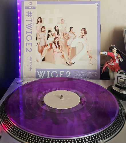 A transparent purpl vinyl record sits on a turntable. Behind the turntable, a vinyl album outer sleeve is displayed. The front cover shows the 9 members of TWICE standing and sitting around a large hashtag and number 2. 

To the right of the album cover is an anime figure of Yuki Morikawa singing in to a microphone and holding her arm out.