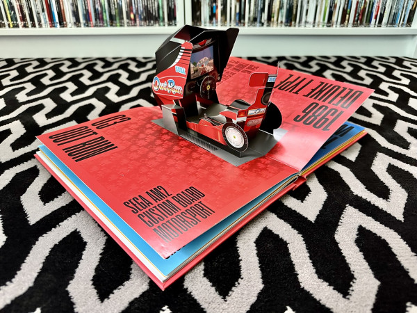 Photo of a large book with a pop up cardboard Outrun arcade machine. It’s a driving seat with a steering wheel and a screen built to look like a car. 