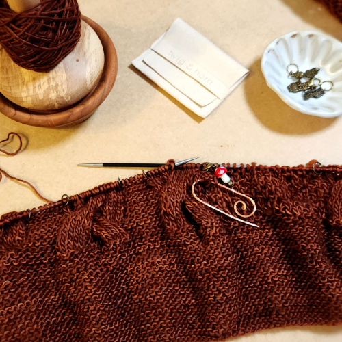 A variegated warm brown knitting project. It has a large section of garter stitch and the current section is large cables with a loose stitch in between. 
Six stitches are being held on a hammered copper cable needle, there is a small mushroom stitch marker.