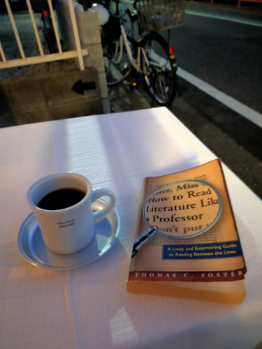 The photo is of an outside white table The light brown paperback book has the title enhanced by a magnifying glass. To the left is a white mug of black coffee with the tiny words COFFEE HOUSE TAKIZAWAKE on it. It is on a round silver metal coaster. A parked whire bicycle can be seen in the distance along the street