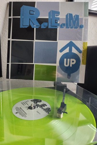 R.E.M. - Up album cover. R.E.M. in blue at the top with squares all over the cover. Arrow pointing up logo on the corner. Vinyl record is green.