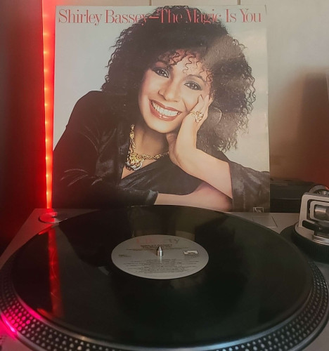 A black vinyl record sits on a turntable. Behind the turntable, a vinyl album outer sleeve is displayed. The front cover shows Shirley Bassey resting her head against her hand as she looks and smiles at the camera. 