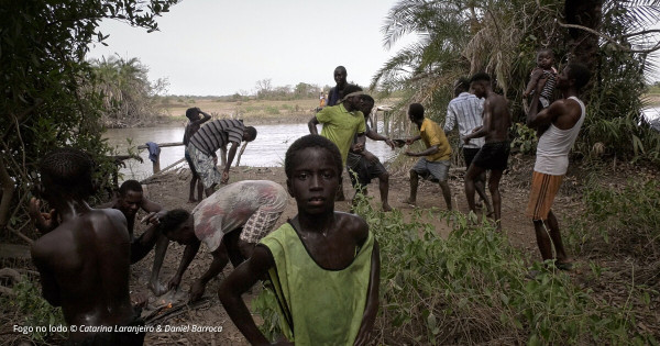 Still from the film "Fogo no Lodo" by Catarina Laranjeiro and Daniel Barroca, showing a group of black boys and adult men by a river. In the foreground, a boy looks directly at the camera.