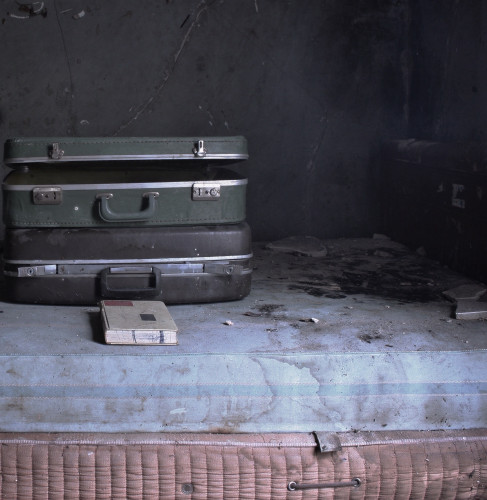 A bedroom in an old, abandoned house. A bed, a few suitcases, & a book are pictured.