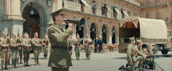 A man in a military uniform speaking through a bullhorn in a town square, surrounded by soldiers with guns