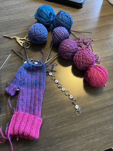 Six tiny balls of yarn in a gradient from pink to blue next to a half knitted fingerless mitten, pink on top and purple-blue at the bottom