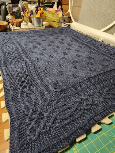 A blue knitted blanket spread out on a table with blocking mats. The inside is a checkered type pattern. Outside is a border with a cable pattern.