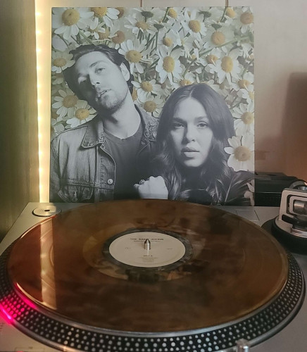 Image shows a turntable with a transparent amber vinyl record on the platter. Behind the turntable vinyl album outer sleeve is displayed. The front cover shows a man and woman looking at the camera surrounded by daisies