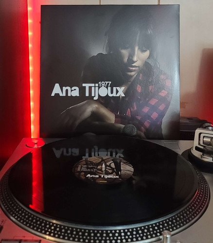 Image shows a turntable with a black vinyl record on the platter. Behind the turntable vinyl album outer sleeve is displayed. The front cover shows Ana Tijoux wearing a flannel shirt while looking down. She has her arms crossed and resting on her leg. One hand is holding a microphone