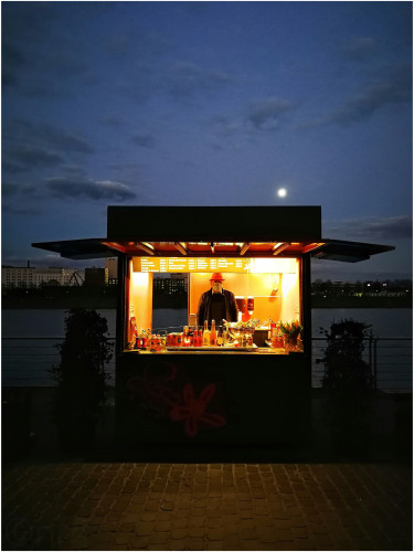 Lonesome masked vendor in a 'cocktail hut' near the river, under a full moon illuminating the dark blue sky and the empty square around his shop.