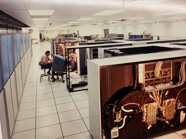 Color photograph. In the foreground is one of the parallel units of the massive ILLIAC IV as it is being installed in a raised floor computer room at NASA Ames Research Center in 1972. In the middle of the photo in the distance of the large room a man sits on a chair at a wheeled workstation working on the installation and there is a partial view of another individual. The room has white ceilings and large white floor tiles.