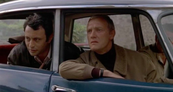 Two men lean out of a little car, watching something intently