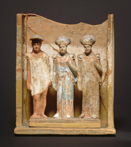 A small terracotta naiskos (shrine or temple, usually funerary), with three figurines facing forward. Hermes, on the left wears a dangerously short white unbelted chiton with a large shield fibula (pin) holding the garment together at his right shoulder. Two other figures are identical women, each wearing a colorful peplos, which reach to the ground. It seems to be striped in red, blue and possibly white pigments. They each wear large upside-down conical hats (like Guinan in Star Trek: The Next Generation TV series) and they have shield fibulae at each shoulder, ho,ding up their garments. All three figures stand on a stepped platform within the shrine, which is missing its pediment.

5th c. BCE, from Tanagra, Boeotia, Greece
Altes Museum, Berlin (TC 6678)
