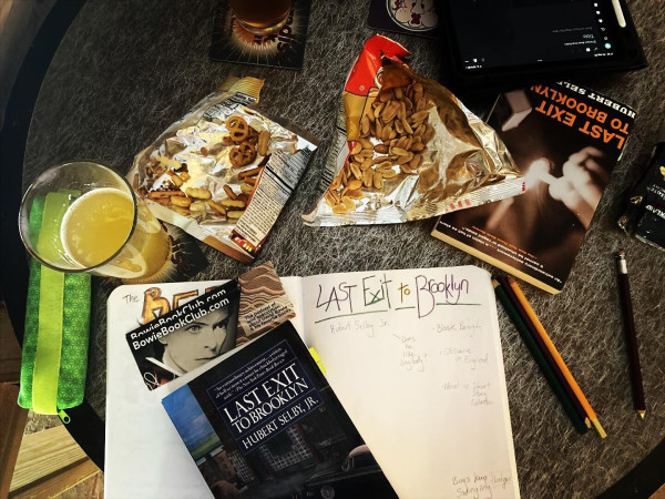 Table top with two copies of the book "Last Exit to Brooklyn" plus notebooks, pens, pints of beer and open snacks.