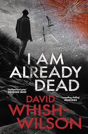 Image of the book cover for I Am Already Dead by David Whish-Wilson. With the quotes 'intelligent and gutsy' - Weekend West and 'compelling, thrilling' Readings, the cover is a dark blacks and grey coloured image of a man standing on the edge of steep sand hill with some scrubby grass beside him. There are seagulls flying overhead and the image is covered by cracked glass, with an obvious damage point at the top right.