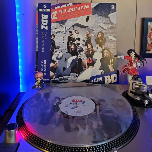 A clear vinyl record sits on a turntable. Behind the turntable, a vinyl album outer sleeve is displayed. The front cover shows the 9 members of TWICE sitting and standing around a damaged wall with the letters B D Z, and pieces of concrete flying around. 