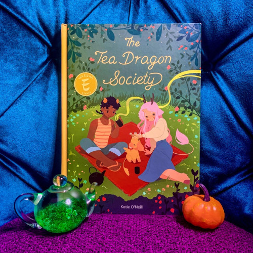 The graphic Novel The Tea Dragon Society by K. O’Neill with a plush royal blue background, purple foreground. And a cute little pumpkin and glass teapot with shamrocks in front of the book.