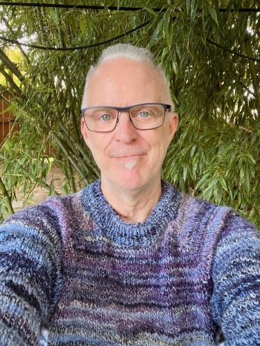 A white man with silver hair, blue eyes and glasses poses in front of bamboo wearing a hand knit sweater, the colors of periwinkle, purple, black, and gray forming narrow stripes. 
