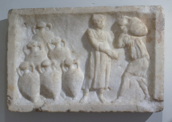 Description from the museum: “The relief appears to show a merchant receiving a delivery from a porter at right, who is carrying a jar over his shoulder. Other amphorae, with their mouths clearly sealed, are stacked in the corner at left.”