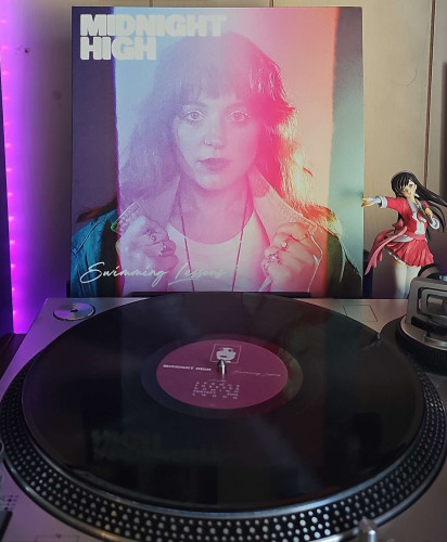 A black vinyl record sits on a turntable. Behind the turntable, a vinyl album outer sleeve is displayed. The front cover shows a woman holding her collar out on her jean jacket. 

To the right of the album cover is an anime figure of Yuki Morikawa singing in to a microphone and holding her arm out.