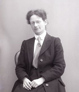 Maud Joachim in 1910 black and white photo seated white woman with dark hair drawn back wire rim glasses wearing dark coat and skirt over white shirt with a mid shade tie with a square pin.