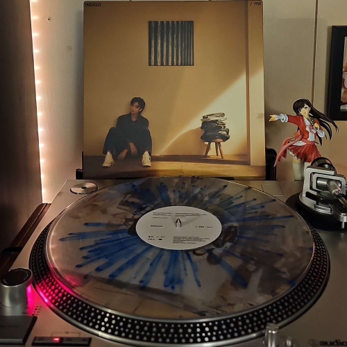 A clear with blue splatter vinyl record sits on a turntable. Behind the turntable, a vinyl album outer sleeve is displayed. The front cover shows RM sitting in a room with clothes stacked on a stool. 