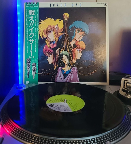 A black vinyl record sits on a turntable. Behind the turntable, a vinyl album outer sleeve is displayed. The front cover shows Big Gold's head which has its hair extending down like roots/tendrils. On the left side of Big Gold are Iczer-1 and Iczer-2, and on the right are Nagisa and Sepia. At the bottom of the hair is Sir Violet.