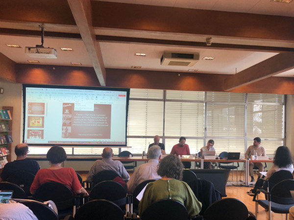 Photograph of the conference room during the panel "Transits". It shows the presentation on the screen, the members of the panel sitting on a table and the audience's backs.