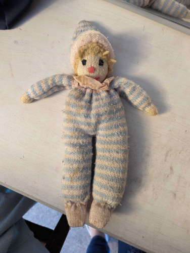 A somewhat ratty old knitted doll, lying on a background of white painted wood. The doll is wearing a striped baby blue and white onesie and matching hat. It has curly blonde hair and a ribbon scarf. It is clearly very old and loved, but very worse for wear.