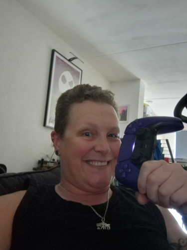 A smiling person (me) holding a purple PS5 controller and looking at the camera. Wearing a black top and a hieroglyphic pendant. 