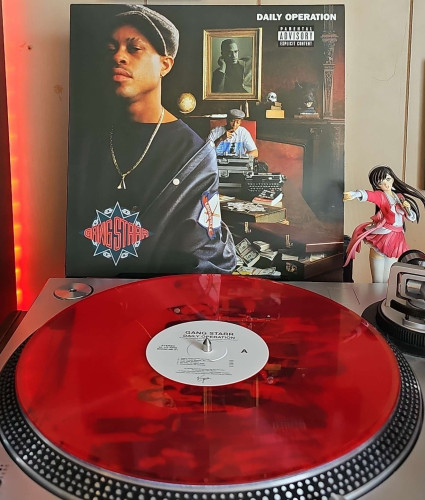 A Red Translucent vinyl record sits on a turntable. Behind the turntable, a vinyl album outer sleeve is displayed. The front cover shows Guru standing close to the camera. Behind him is DJ Premier sitting in a chair smoking a cigar with a trunk in front of him.. 

To the right of the album cover is an anime figure of Yuki Morikawa singing in to a microphone and holding her arm out. 