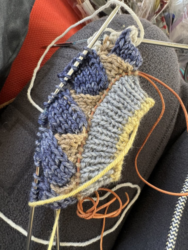 Entrelac knitting. Dark blue rectangles coming off brown triangles attached to light blue ribbing and yellow cast on