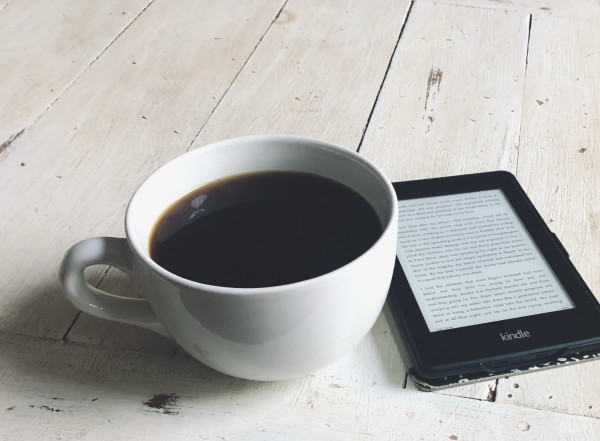 A large cup of coffee sitting next to my Kindle on a wooden table.