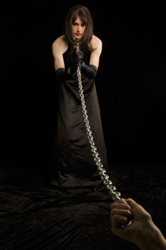 A crossdresser in a black satin gown with wrist shackles and a leash