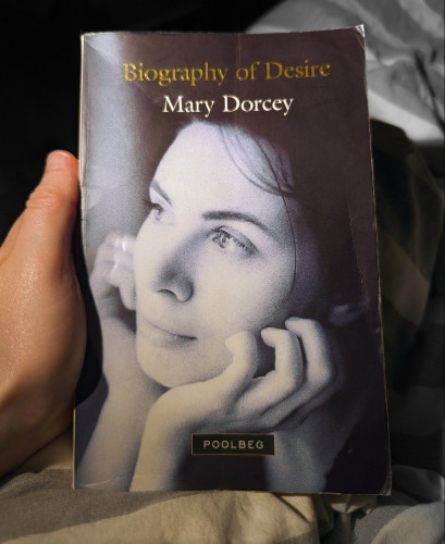 Book cover of Mary Dorcey's Biography of Desire that shows a causcasian woman holding her head in her hands looking pensive. Black and white. Poolbeg press publishing branding at the bottom of the cover.