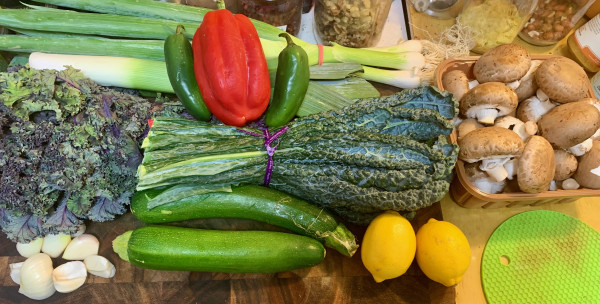On  a cutting board: a leek, a bunch  of young onions, a red bell pepper, two  jalapeños, a bunch of red kale, a bunch of Tuscan (dino) kale, two green zucchini, about 8 large cloves garlic, 2 lemons, and a big box of medium-sized crimini mushrooms