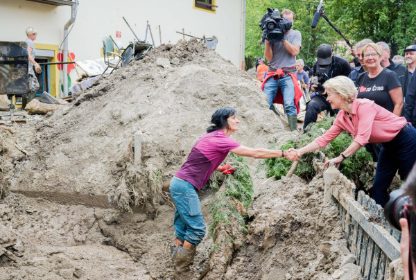 President Von der Leyen shaking hands with a Slovenian citizen, who is standing on the grounds where the floods happened, surrounded by a big pile of sand.