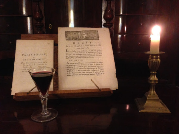 Two contemporaneous publications on the outbreak of the Revolution on a wooden reading stand on the desktop of a Biedermeier secretary.

Left foreground, a small glass with red wine;
Right: a candle in a brass candlestick