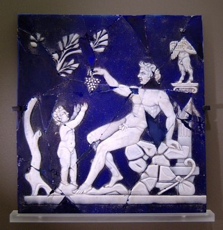 A satyr is holding a cluster of grapes with the child Dionysos reaching out his little hands towards it. The scene is framed by a tree on the left, behind little Dionysos, and a small statue of Pan atop a column on the right, behind the satyr. The scene is depicted in white against a blue glass background.