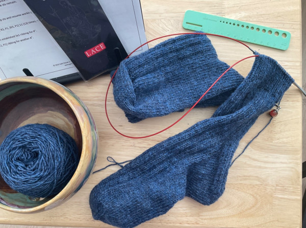 Hand knit sock in progress in blue wool on circular needle with red cable. Folded completed sock sits under cable beside green ruler, yarn bowl and yarn on light wood table. 