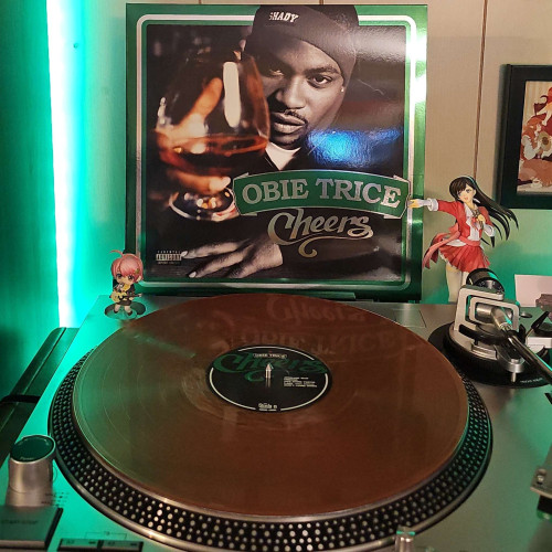 A Cognac vinyl record sits on a turntable. Behind the turntable, a vinyl album outer sleeve is displayed. The front cover shows Obie Trice holding out a snifter glass filled with alcohol. 