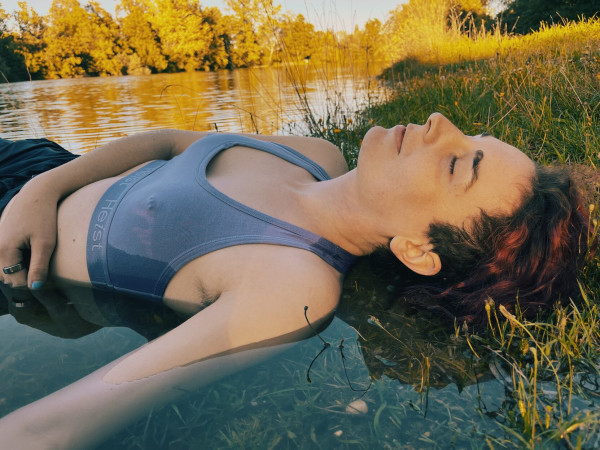 Zora, a white trans woman in her 20s with red curls, lying half in the water and half among grass and wildflowers at the lakeshore. She's giving soft futch, made even softer by the golden hour light on her face. Nipple piercings poke through her bralette. The lake is surrounded by warmly lit trees. 