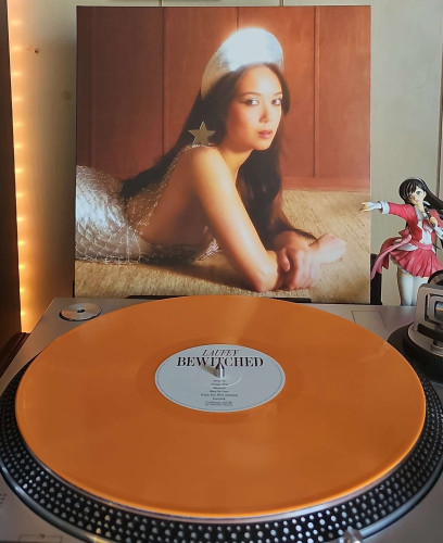 An orange vinyl record sits on a turntable. Behind the turntable, a vinyl album outer sleeve is displayed. The front cover shows Laufey laying on her stomach on the floor. She is wearing a silver headpiece, dress and star earring. 

To the right of the album cover is an anime figure of Yuki Morikawa singing in to a microphone and holding her arm out. 