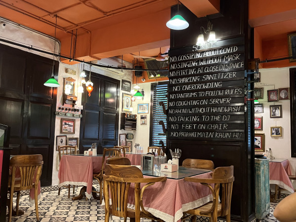 The interior of Sodabottleopenerwalla in Khan Market. The restaurant features wooden tables with red checked table clothes, old antique wall hangings and photographs. There is a large sign of rules in the center. The rules a quite tongue-in-cheek, including "No gossiping about Covid, " No singing without mask" "No tantrums to follow rules." 
