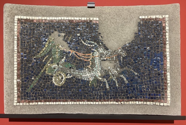 Description from the museum: “Originally located on a wall treated with pumice stone and stucco and punctuated by pilasters, this concave mosaic was meant to decorate one of the semicircular niches present in the wall. At the centre, a chariot can be seen pulled by two fawns with the rear part of their bodies in the shape of fish. A highly deteriorated figure, which can perhaps be identified as a long-tailed parrot, is driving the chariot.”